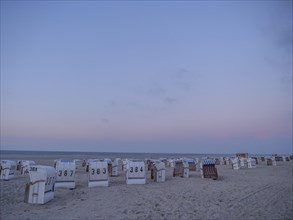 A quiet beach with white and striped beach chairs at dusk under a pastel-coloured sky, setting sun