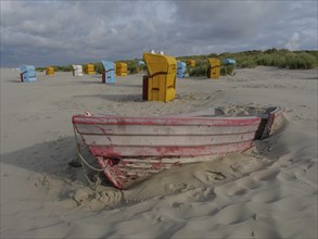 Old, half buried boat in front of colourful beach chairs on sandy beach, cloudy sky and dunes,