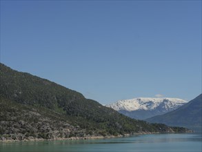 Natural landscape with calm lake, snow-capped mountains and sunny sky, greenish shimmering water in