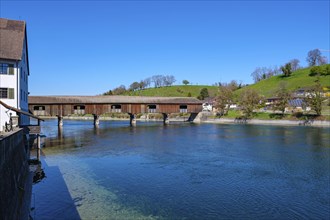 Wooden bridge over a river with old buildings in the background and a green meadow under a bright