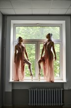 Two graceful ballerinas in transparent long dresses stand on the windowsill in the corridor