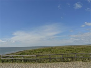 View of the sea and a meadow with a wooden fence under a partly cloudy sky, dune and beach by the