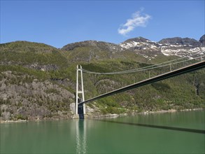 Massive bridge over a green lake, surrounded by forested and rocky mountains under a clear blue