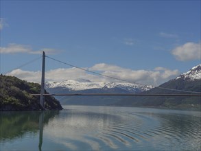 A suspension bridge stretches over calm water surrounded by snow-capped mountains, bridge in a