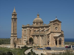 Neo-Romanesque basilica made of light-coloured stones with a high bell tower and some cars in the