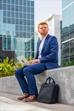 Vertical full length photo of a distracted businessman sitting outside a financial building using