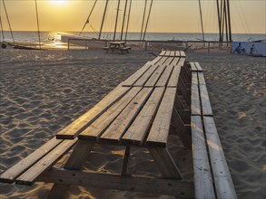Long picnic tables on sandy beach at sunset, boats in the background, warm and relaxing atmosphere,