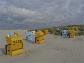 Row of colourful beach chairs standing in the sand, surrounded by dunes and a cloudy sky, colourful