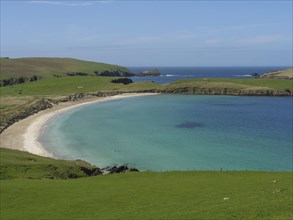 Calm bay with golden sandy beach and blue water, surrounded by green grassland and a clear sky,