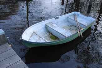 An abandoned rowing boat floats next to a jetty on calm water, surrounded by spring atmosphere and
