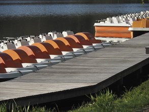 Wooden jetty with several orange and white pedal boats on a peaceful lake, rowing boats and pedal