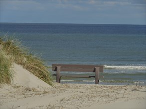 A bench in front of the sand dunes with grass, in front of it the blue sea and a cloudy sky, dunes