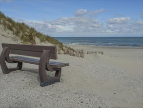 A bench by the sand dunes with grass, behind it the sea and above it a cloudy sky, dunes on an