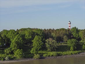 A red and white lighthouse in the middle of dense trees, with clear blue sky and river in the