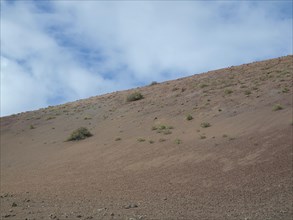 A brown hill with sparse vegetation under a slightly cloudy sky, barren landscape with roads,
