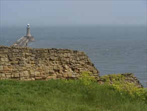 A lighthouse on a pier with old wall remains and green grass in the foreground, ruins and old stone