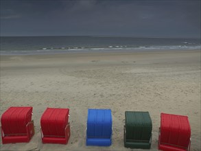 Five colourful beach chairs in red, blue and green stand on a quiet beach in front of a cloudy sky,