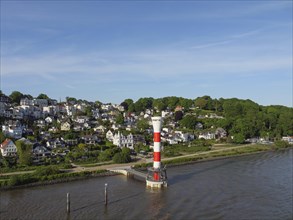 A lighthouse on the riverbank in a green landscape with hills and houses in the background under a
