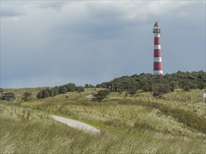 A red and white striped lighthouse in a grassy and wooded dune landscape under a cloudy sky, red
