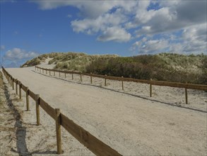 A sandy path lined with a wooden fence, sandy dunes and a sky with clouds, dunes on an island with
