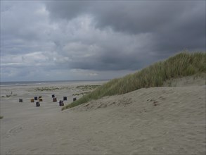 A coastal landscape with cloudy sky, sand dunes and several colourful beach chairs on the beach,