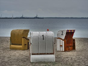 Three beach chairs on a sandy beach in front of the calm sea, ships on the horizon under a cloudy