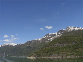 A clear day with snow-capped mountains under a blue sky with scattered clouds, bridge in a fjord