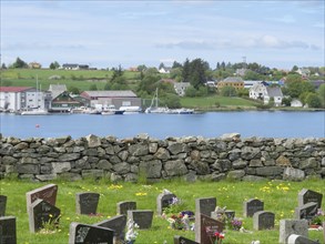 Cemetery with many gravestones, behind it a lake with boats and houses on the hill, under a