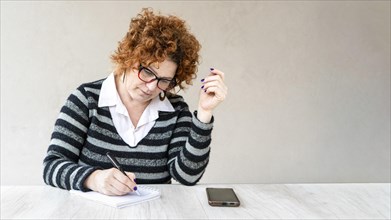 A woman with red hair and glasses is writing in a notebook. She has a cell phone on the table in