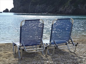 Two blue deckchairs on the beach with a view of calm sea and rocks in the background, sun umbrellas