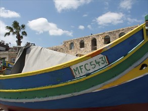 A colourful wooden boat in the harbour with buildings and palm trees in the background, many