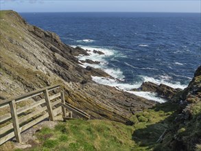 Rocky coastline with a wooden staircase leading to the stony cliffs and the blue sea, calm sea with