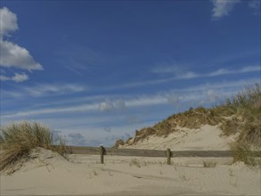 Sand dunes on the beach under a clear blue sky with scattered clouds and tufts of grass, beach and