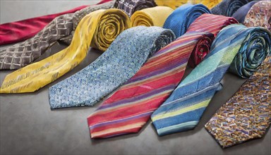 Several colourful ties, partly rolled up, with different patterns and textures on a grey surface,