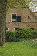 Brick house with black shutters and numbered facade, surrounded by a green meadow and flowers, old