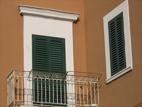 A balcony in front of an orange-coloured wall, decorated with green shutters and a simple railing,
