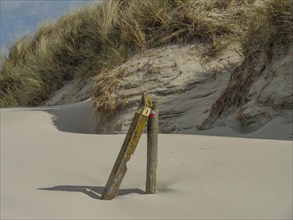 Wooden post in a sand dune with grasses, under a clear sky with few clouds, dune and beach by the