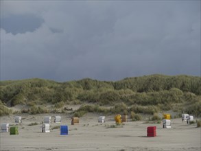Beach chairs are spread out in front of a hilly dune landscape under a cloudy sky, colourful beach