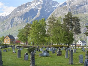 Green cemetery with gravestones in front of an impressive mountain scenery and a house in the