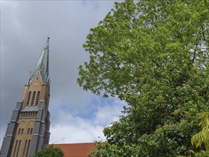 A tall gothic church tower next to a lush green tree against a cloudy sky, large church towers with