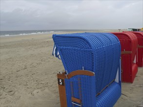 A blue beach chair stands on the beach with other red beach chairs in the background under a cloudy