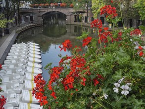 River with white boats on one side, red flowers in the foreground and a stone bridge in the