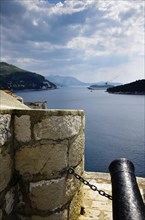 View of the sea with stone coast, chain and cannon in the foreground, hills and cloudy sky in the