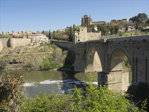 Stone bridge with arches over a river, surrounded by a cityscape and vegetation, sunny weather,