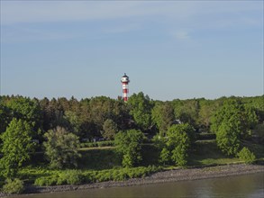 A red and white lighthouse stands in the middle of trees, with the river in front of it and a