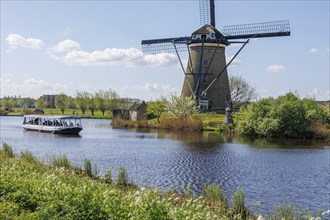 A windmill next to a river through which a boat is sailing on a beautiful spring day, many historic