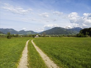 Field path leads into a meadow with hay barn, snow-covered mountain peaks in the background, near