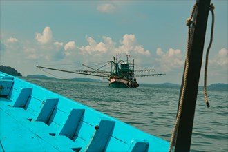 A fishing boat is seen on calm ocean waters under a cloudy sky from the bow of a blue boat, showing