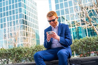 Confident businessman smiling while using phone sitting in the city next to financial buildings