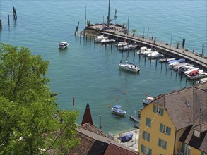 View of a harbour with boats at the pier, behind it houses and a calm blue lake, old buildings with
